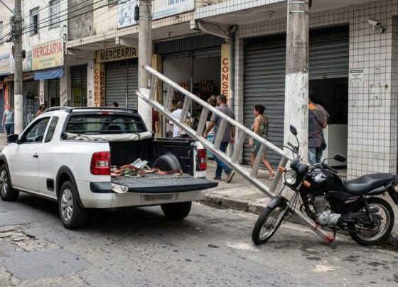 Motorista transporta uma escada presa na caçamba de uma caminhonete, perde o objeto em uma curva e danifica uma moto estacionada, mas segue viagem sem perceber, enquanto a câmera de uma loja registra a queda e a identificação do veículo
